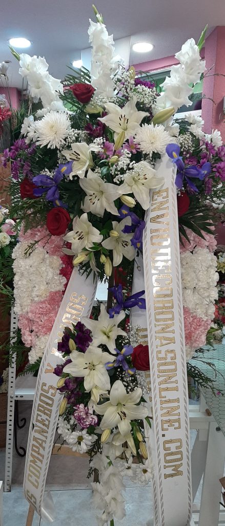 Corona funeraria doble cabecero esta compuesta de gladiolos liliun blanco, anastasias blancas, iris azules y rosas rojas, terminada en dos brazos de clavel blanco y rosa